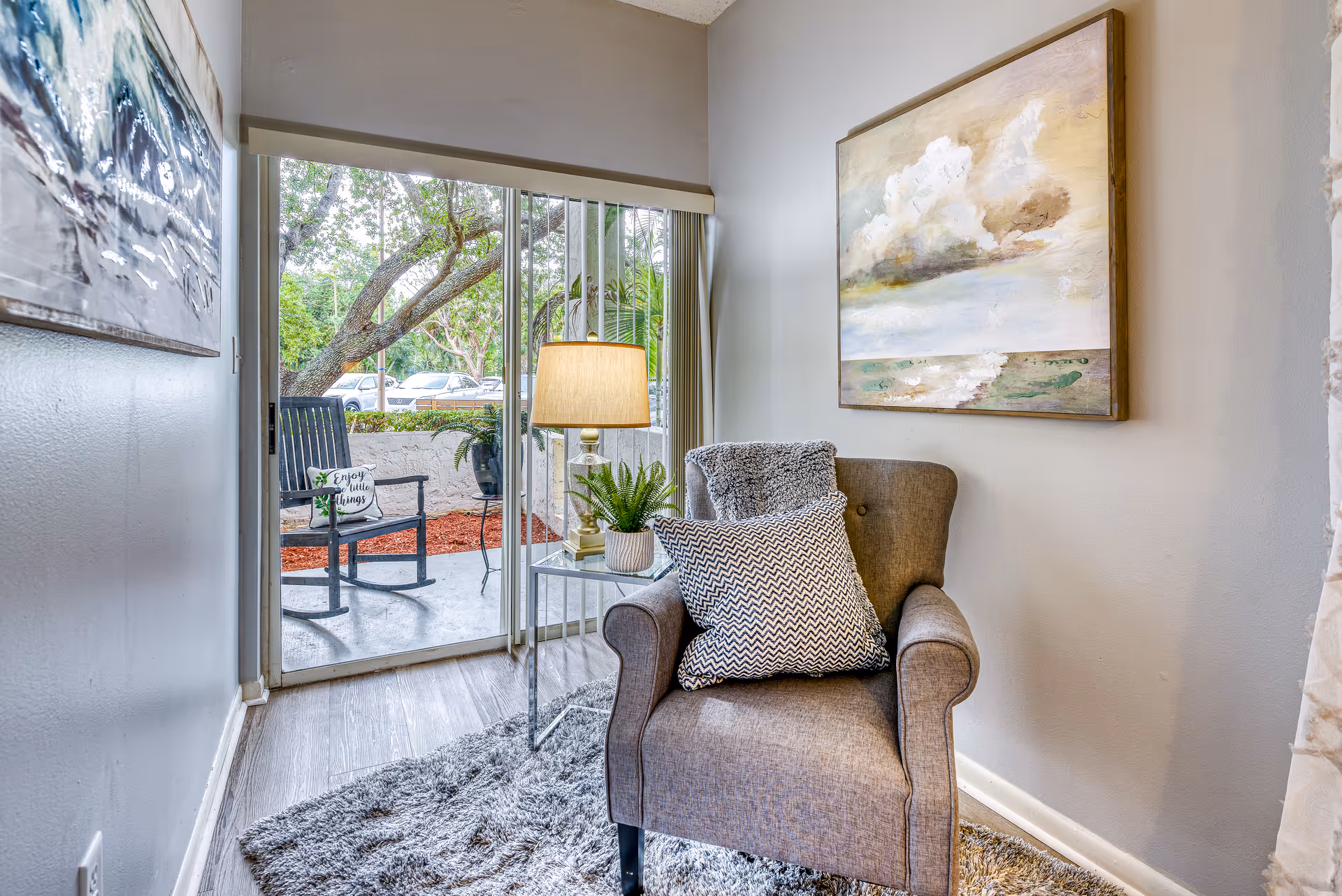 A cozy corner in a senior living facility featuring a comfortable armchair with a patterned pillow and a throw blanket. Next to the chair is a small glass side table with a lamp and a potted plant. The wall behind the chair has a large painting of a cloudy sky over a landscape. Sliding glass doors open to a small outdoor patio with a black rocking chair and a cushion that reads 'Enjoy the little things'. Trees and parked cars are visible outside.