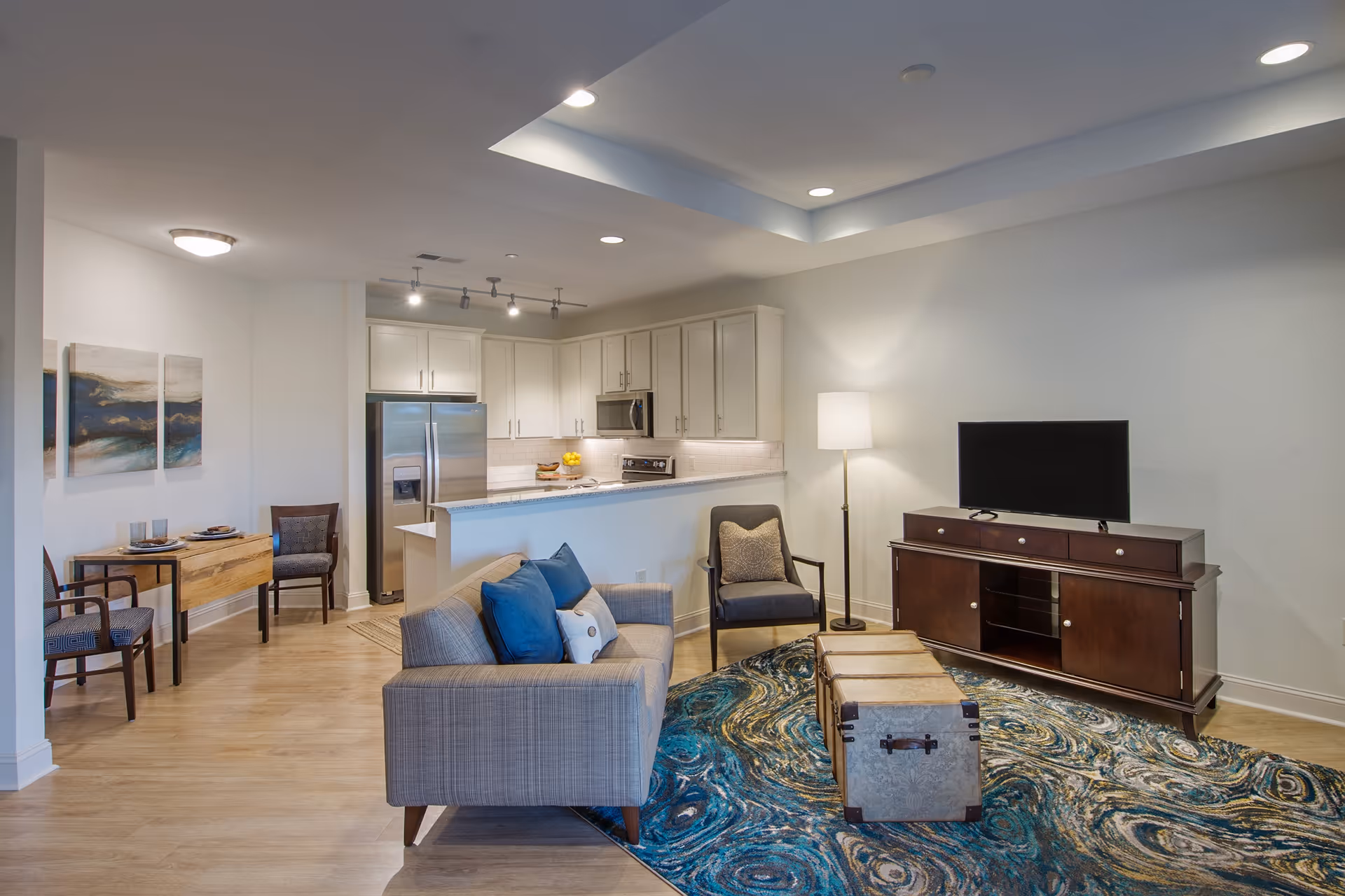 Open-concept living room and kitchen with a sofa and pillows, armchair, TV on a console, patterned rug, and white kitchen cabinets with stainless steel appliances.