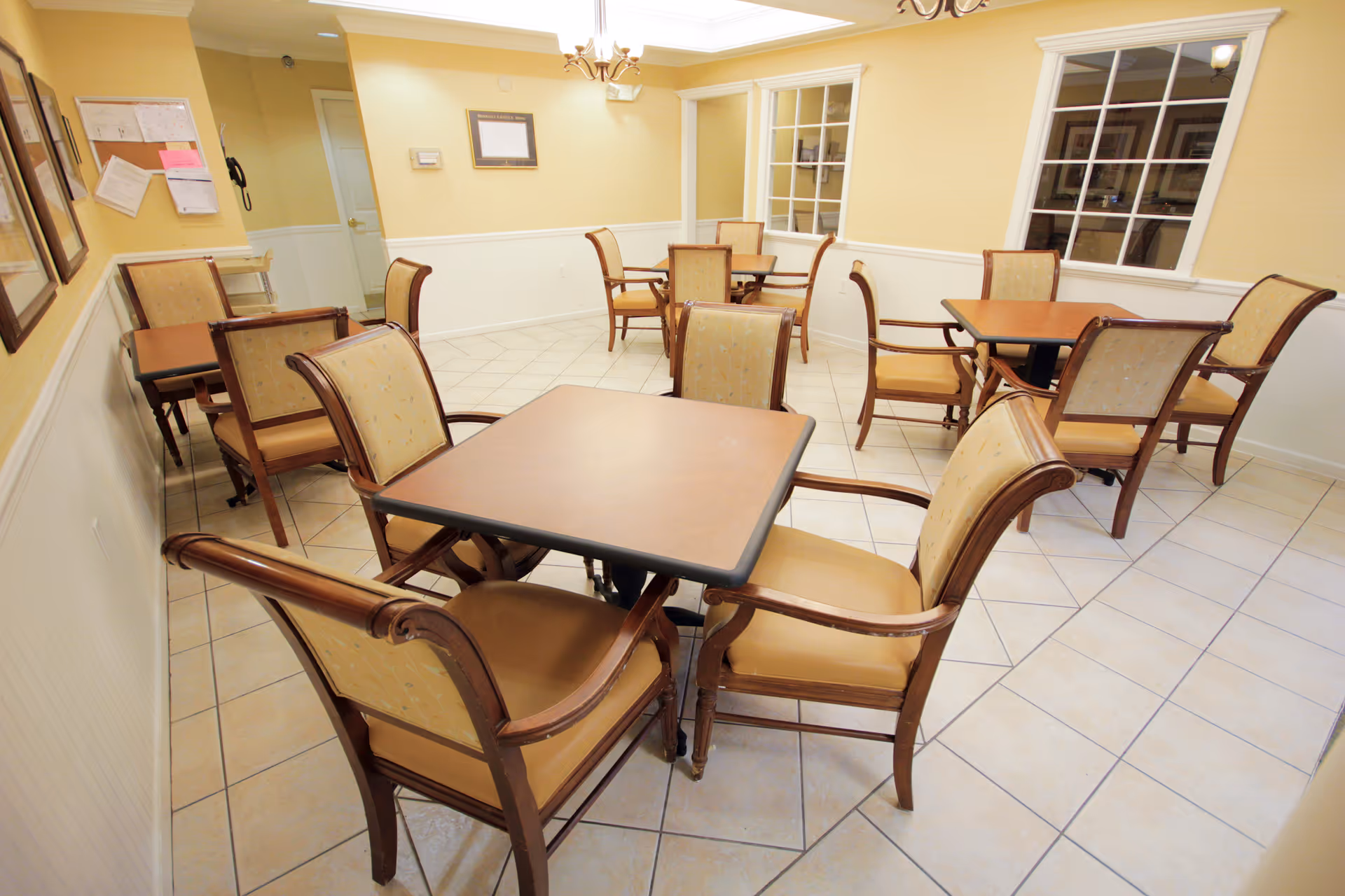 Bright dining room with tiled floor, several square tables and upholstered wooden chairs arranged for residents.
