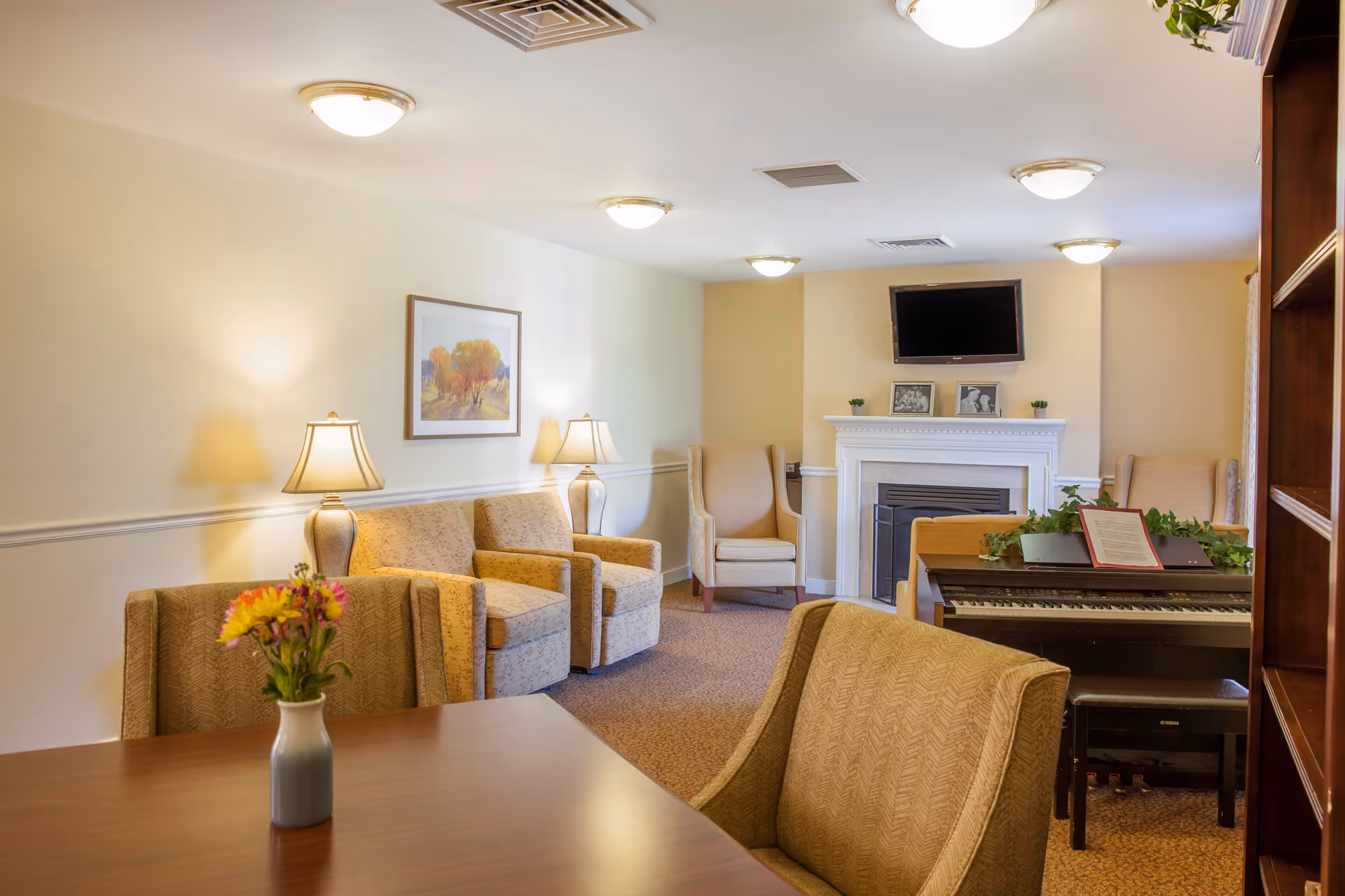 A cozy senior living common room with armchairs, a fireplace with a wall-mounted TV, a piano, and a table with a vase of flowers.