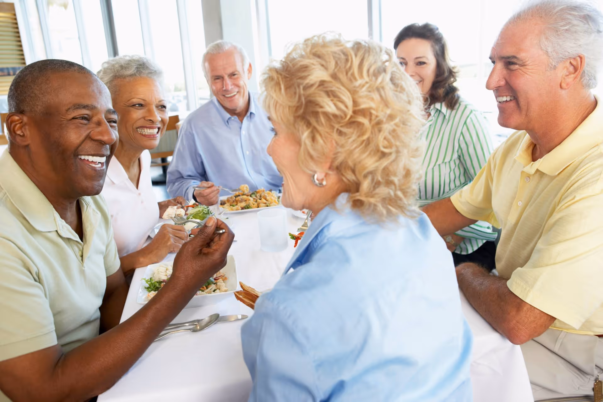 A group of six older adults sitting around a dining table, smiling and enjoying a meal together in a bright room with large windows.