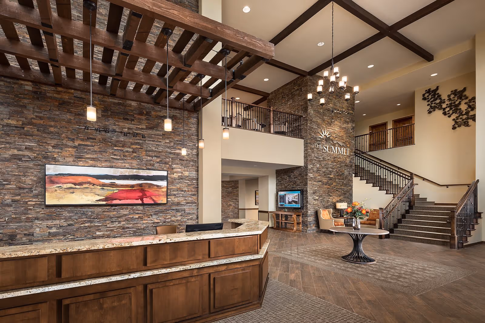 Spacious and elegant lobby area of a senior living facility with a wooden reception desk, stone accent walls, hanging pendant lights, a large abstract painting, a round table with a flower arrangement, and a staircase leading to an upper level. The wall features the text 'THE SUMMIT'.