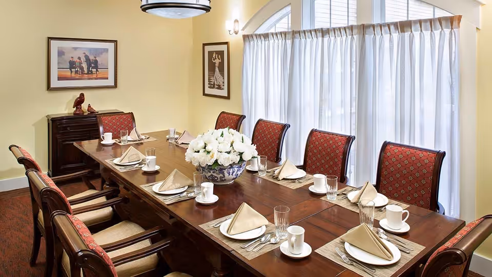 A formal dining room with a long wooden table set for eight people. Each place setting includes a white plate, folded beige napkin, glass, cup, and silverware. The chairs have red patterned upholstery. A large floral centerpiece with white flowers is in the middle of the table. The room has yellow walls, two framed pictures, a small wooden cabinet, and large windows with white curtains allowing natural light.