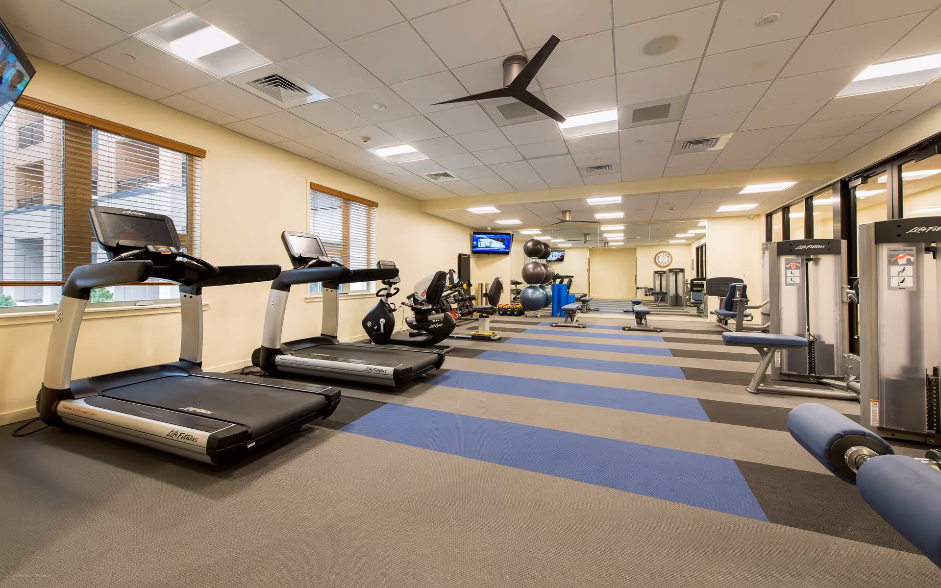 A spacious fitness room with two treadmills, exercise bikes, weight machines, and various exercise balls. The room has large windows with blinds, a ceiling fan, and multiple wall-mounted televisions. The floor is carpeted with a blue and gray pattern.