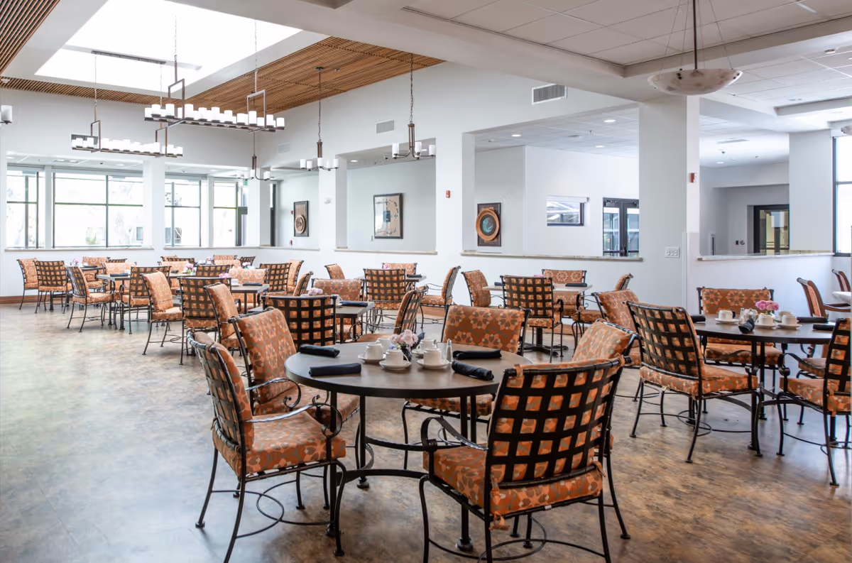 A spacious dining room with multiple round tables and cushioned chairs featuring a patterned orange fabric. The tables are set with white teapots, cups, and black napkins. Large windows allow natural light to fill the room, and modern chandeliers hang from the ceiling with wooden slats. The walls are white with framed artwork.