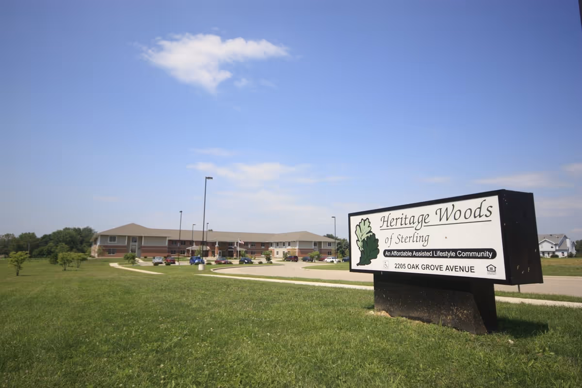 Exterior view of Heritage Woods of Sterling, an assisted living community, showing a large building with a parking lot and a sign in the foreground on a grassy area under a blue sky with some clouds.