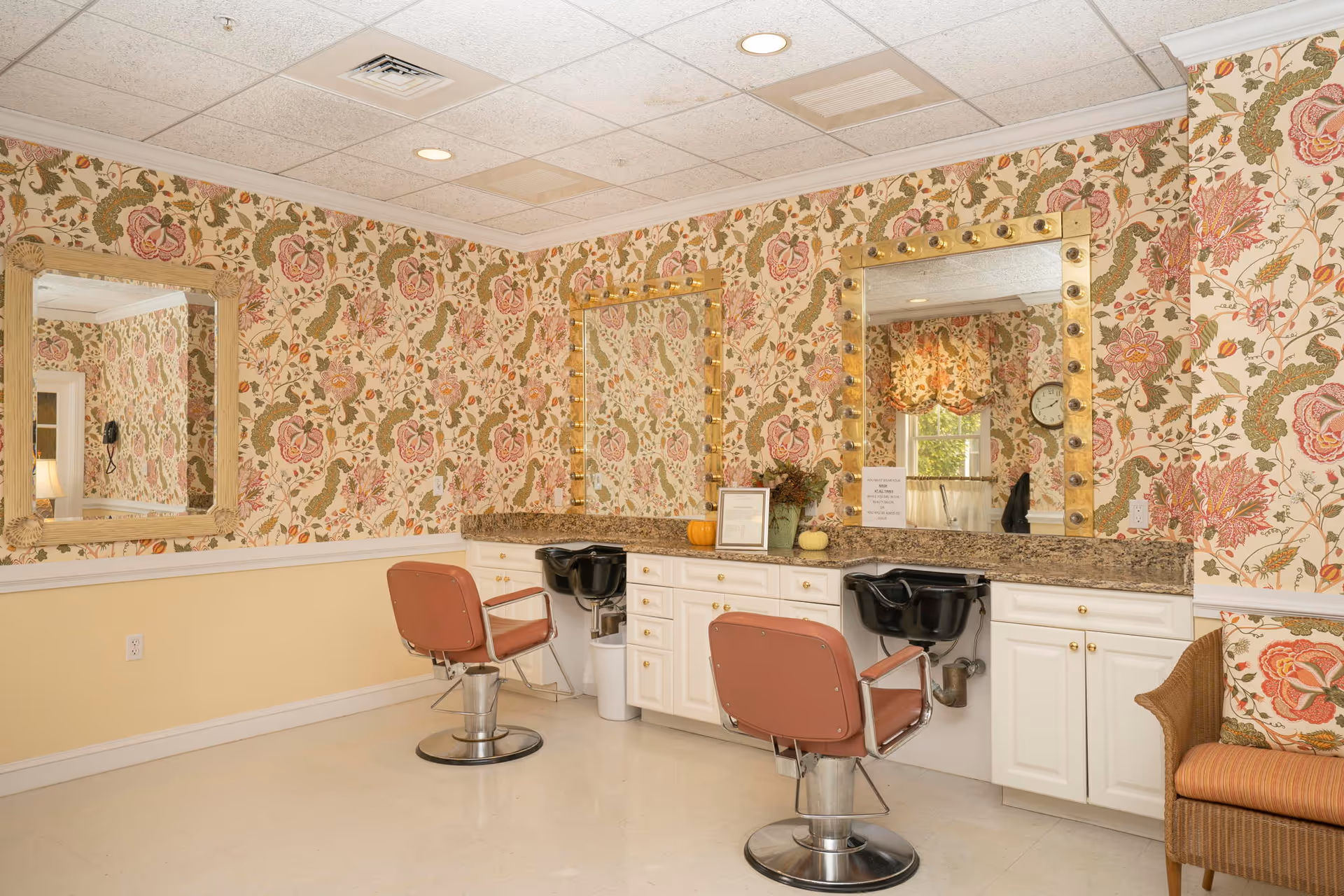 Interior of a salon area in an assisted living community featuring two vintage-style salon chairs in front of a counter with two large mirrors framed with light bulbs. The walls are decorated with floral wallpaper and there is a wicker chair with a floral cushion on the right side.