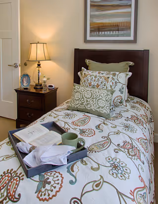 Single bedroom with a patterned twin bed, decorative pillows, a bedside table with a lamp, and a tray holding a cup and book.