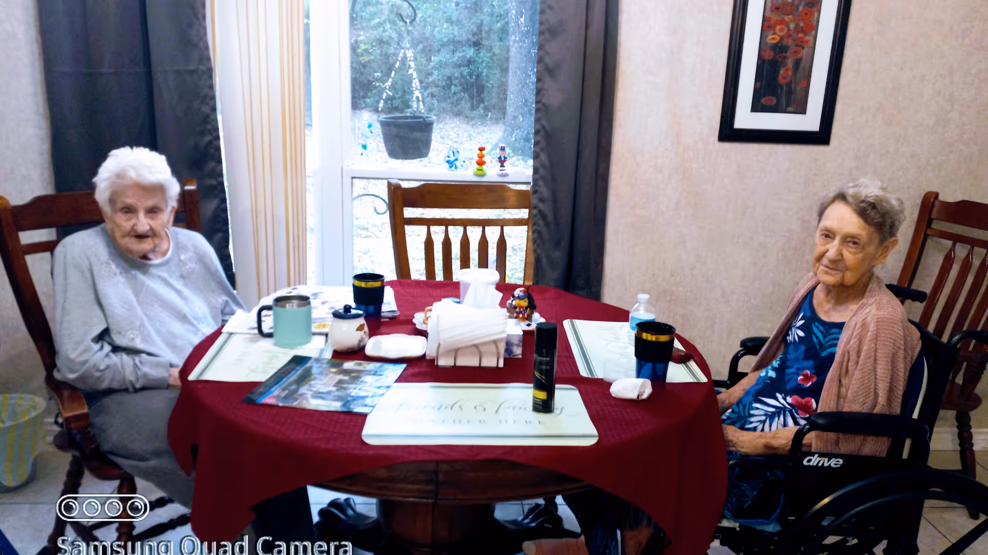Two elderly women seated at a dining table with a red tablecloth in a dining room.