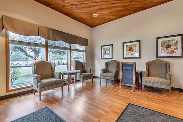 A cozy seating area with four upholstered armchairs arranged around two small wooden tables. The room has large windows with beige valances, allowing natural light to brighten the space. The walls are decorated with three framed floral artworks, and a small chalkboard sign stands near the chairs. The floor is wooden, and the ceiling has a wood panel design.