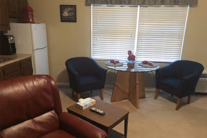 Small living area with a red leather recliner, two navy armchairs around a glass-top table, a side table with a remote and tissues, and a kitchenette with a refrigerator.