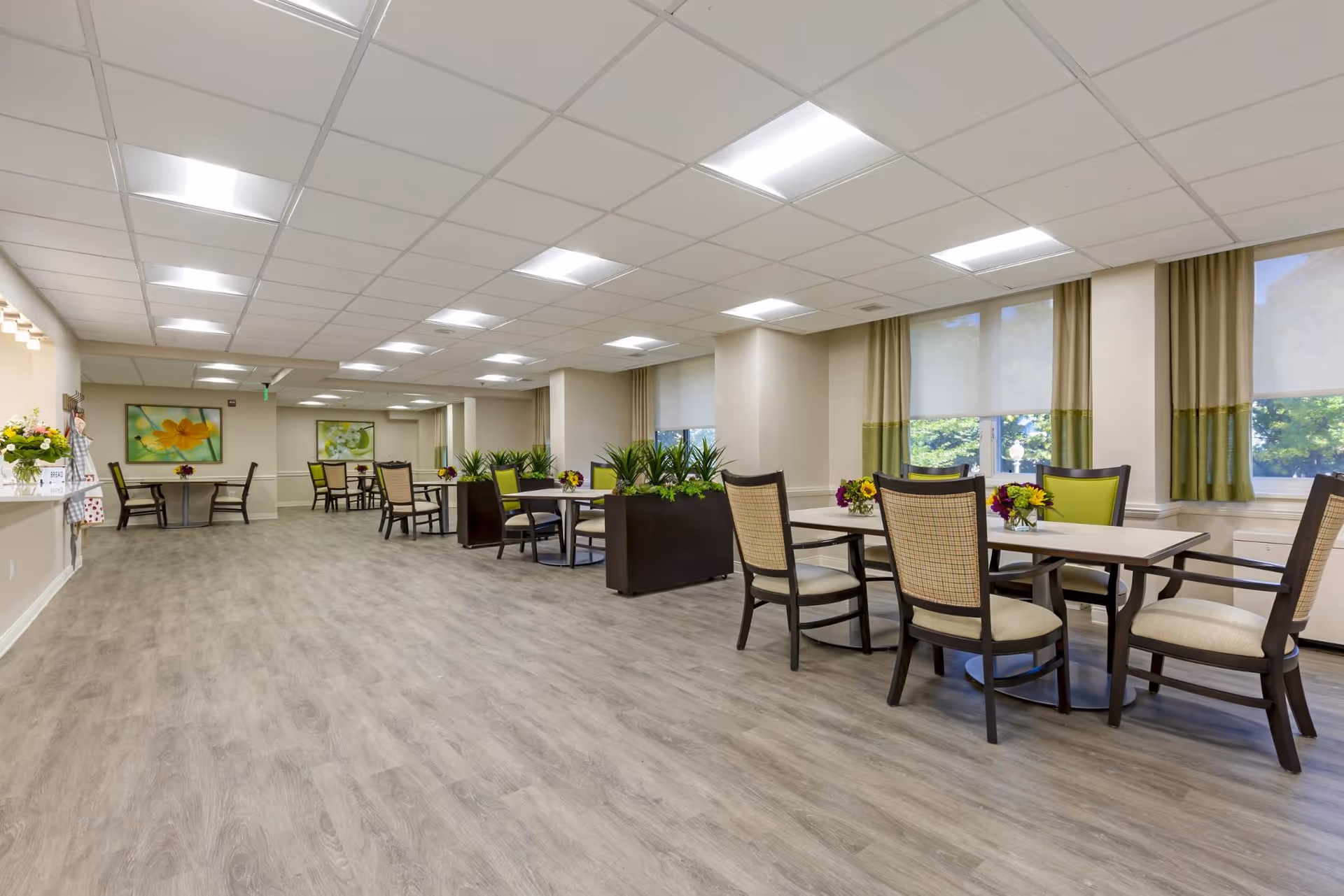 A spacious, well-lit dining area in a retirement community with multiple tables and chairs arranged neatly. The room features large windows with green and beige curtains, potted plants as room dividers, and framed floral artwork on the walls. The floor has a light wood finish, and the ceiling has recessed lighting panels.