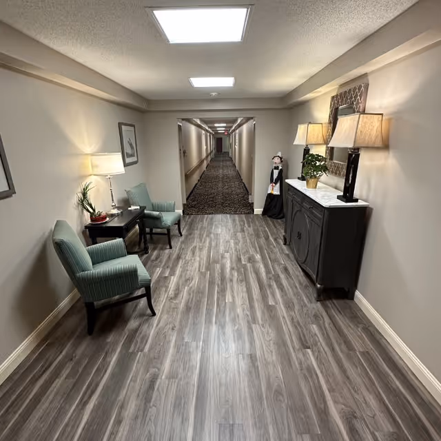 A well-lit hallway in a senior living facility with wood-look flooring and beige walls. On the left side, there are two green upholstered chairs with a small table and lamp between them. On the right side, there is a dark wooden cabinet with two lamps and a potted plant on top, along with a decorative mirror above it. At the end of the hallway, there is a carpeted corridor with multiple doors. A decorative figure stands near the cabinet.
