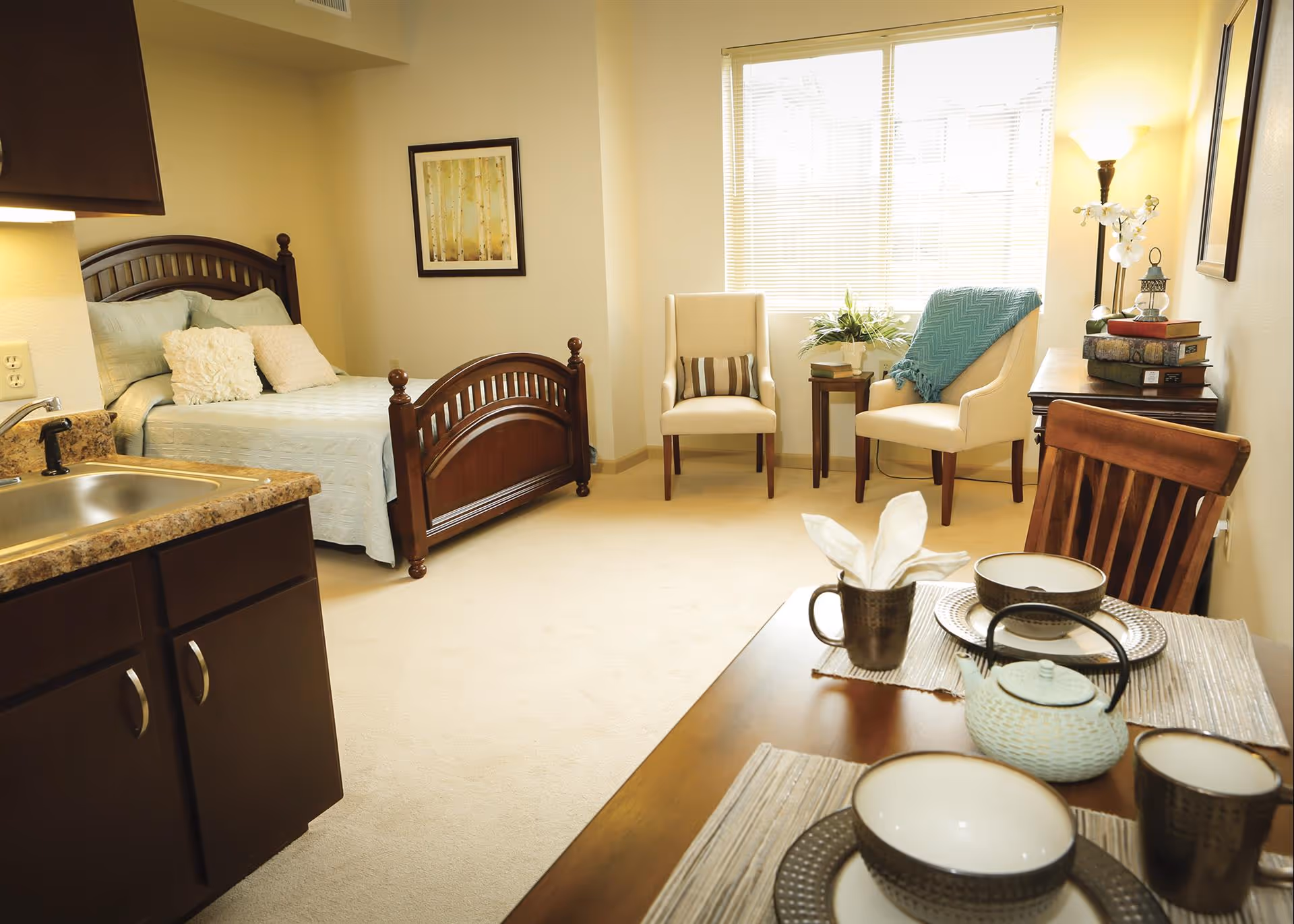 Furnished studio apartment interior featuring a wooden bed, kitchenette sink, dining table set, and seating by a bright window.