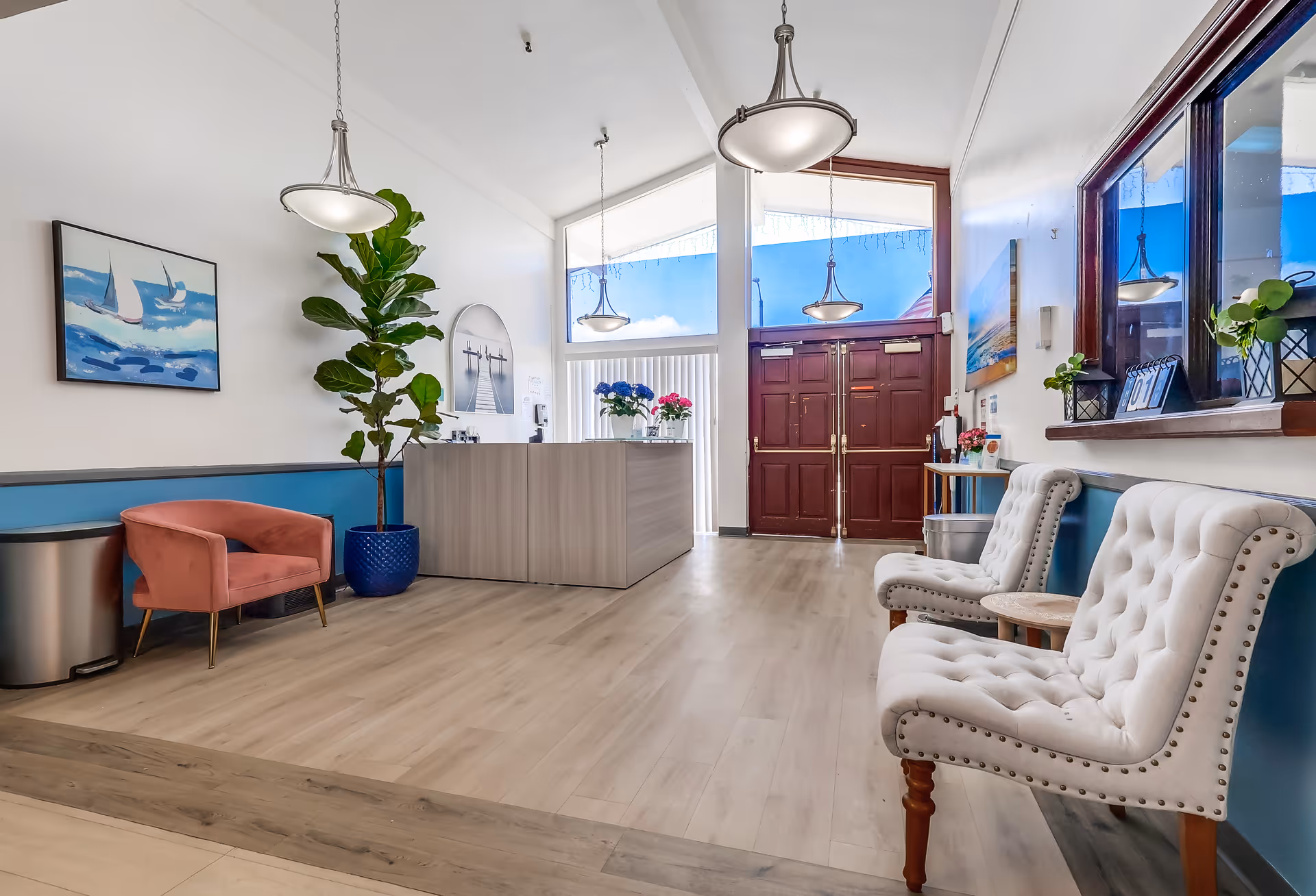 Bright and welcoming reception area with light wood flooring, a modern reception desk, and large double wooden doors with glass panels above. The room features white walls with a blue accent wall, two white tufted chairs with wooden legs, a small round wooden table, a pink armchair, a large potted plant in a blue pot, and hanging pendant lights. Artwork and decorative plants are displayed on the walls and surfaces.