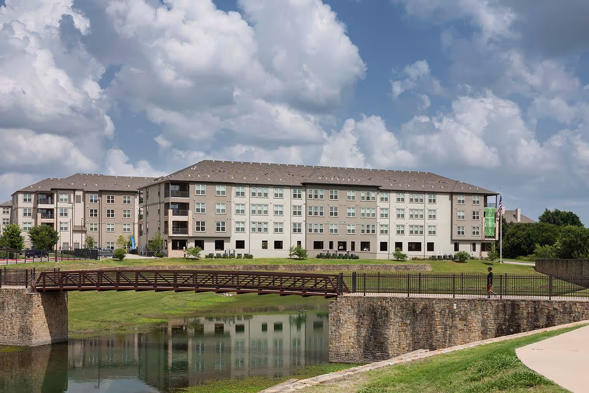 A large multi-story senior living facility building named McDermott Crossing is shown under a partly cloudy sky. In front of the building is a stone and metal pedestrian bridge crossing over a small pond, with a person walking on the pathway near the bridge. The area is landscaped with grass and trees.