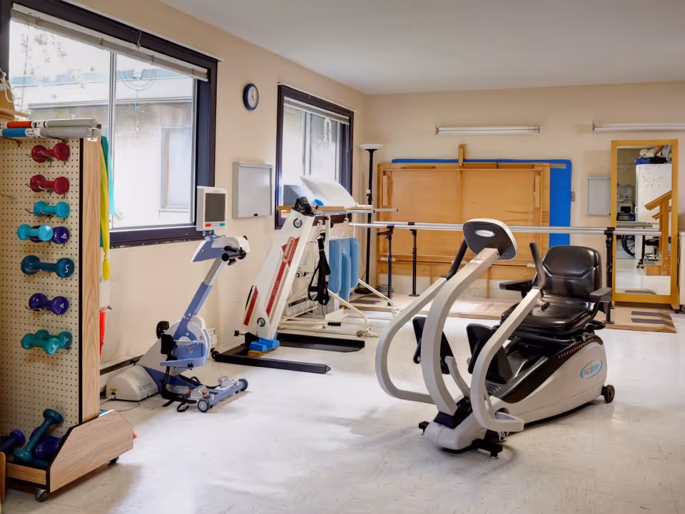 Therapy/exercise room with a recumbent exercise bike, other rehabilitation equipment, a rack of hand weights, and parallel bars.