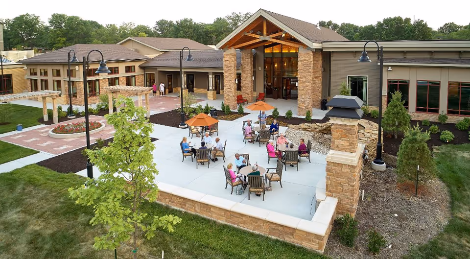 Outdoor patio area at John Knox Village with several elderly people sitting at tables under orange umbrellas, surrounded by a stone wall and landscaped greenery, adjacent to a building with large windows and stone pillars.