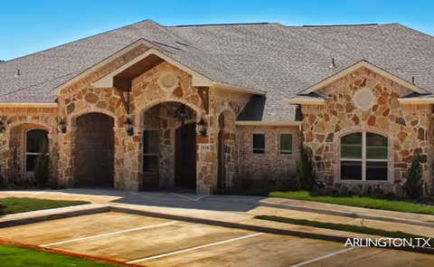 Exterior view of a single-story stone building with a gabled roof and arched entrance, surrounded by a paved parking area and some greenery.