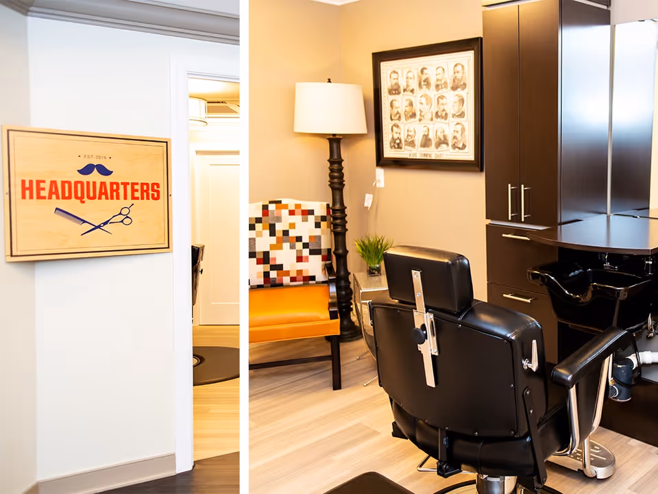 Interior view of a hair salon area in a senior living facility. The image shows a black barber chair facing a black hair washing station with cabinets. To the left, there is a colorful chair with a patchwork design and a tall floor lamp beside it. A framed vintage-style portrait collage hangs on the beige wall. A wooden sign on a white wall outside the room reads 'HEADQUARTERS' with a mustache and scissors graphic.