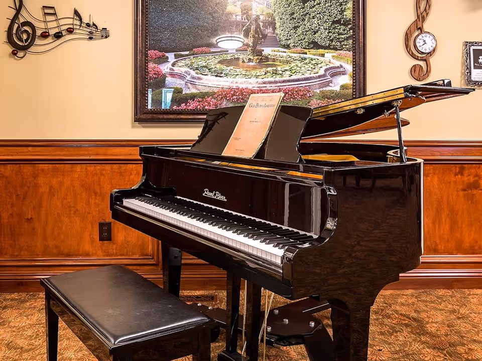 A glossy black grand piano with a bench sits in a wood-paneled common room decorated with framed artwork and musical wall ornaments.