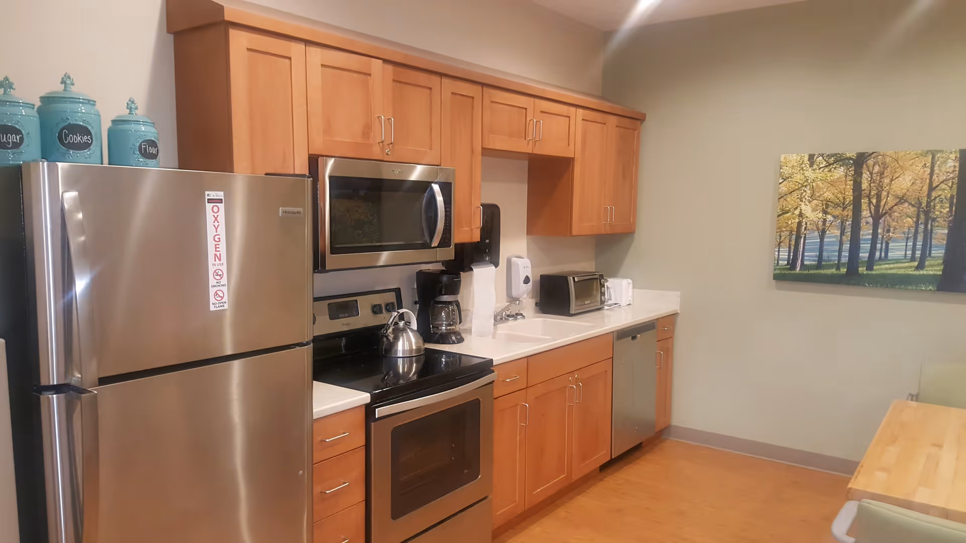 A kitchen area with wooden cabinets, a stainless steel refrigerator with an oxygen use warning sign, a microwave, an electric stove with a kettle, a coffee maker, a toaster oven, a toaster, a sink with a soap dispenser, and a wooden table with chairs. A framed picture of trees is hanging on the wall.