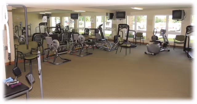 A spacious fitness room with various exercise equipment including stationary bikes, rowing machines, and weight machines. Several green chairs are placed around the room, and large windows allow natural light to fill the space.