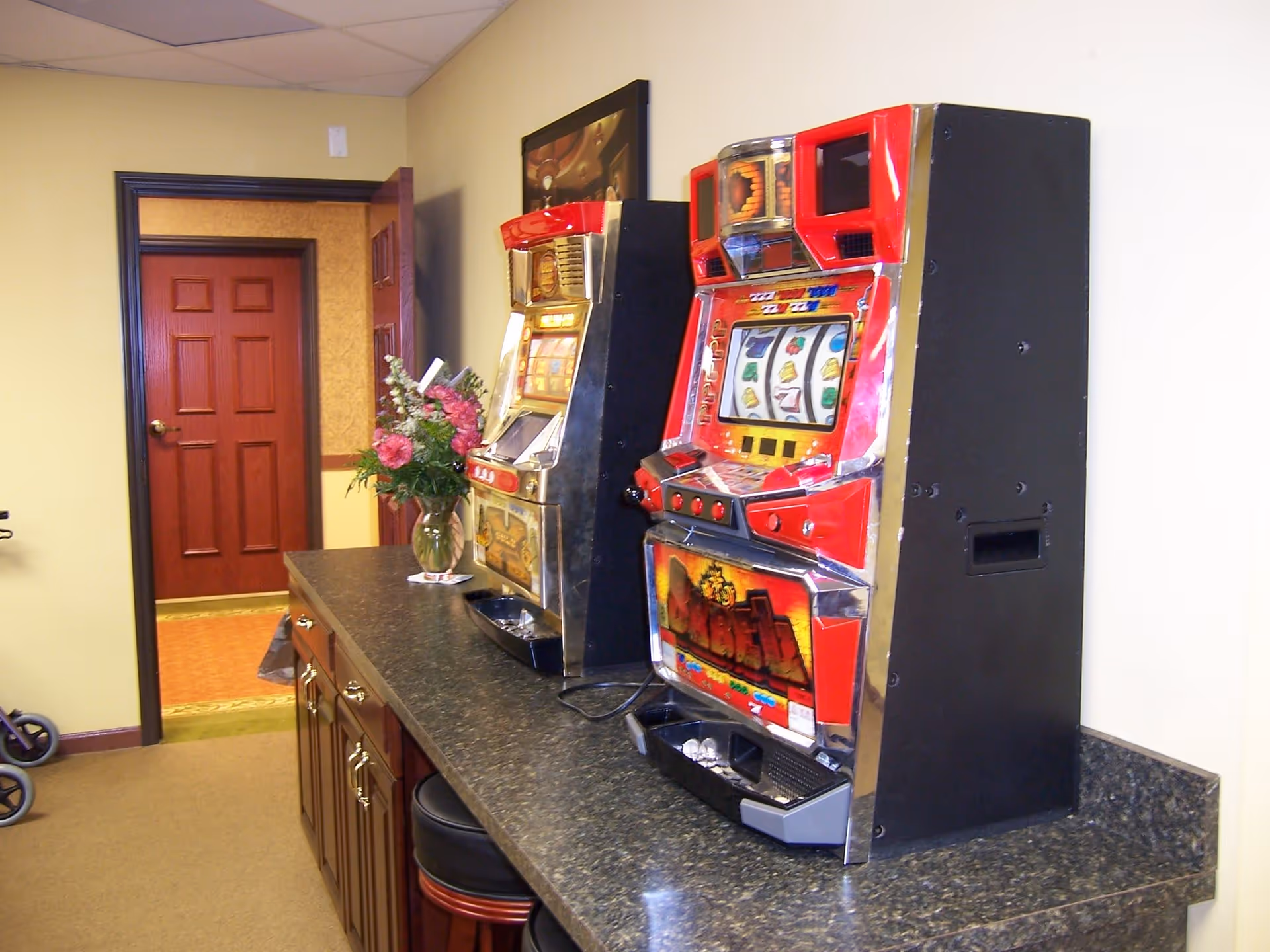 Two slot machines placed on a dark granite countertop in a room with beige walls and carpeted floor. There is a vase with pink and white flowers on the counter, and a wooden door with a brass handle is open in the background.
