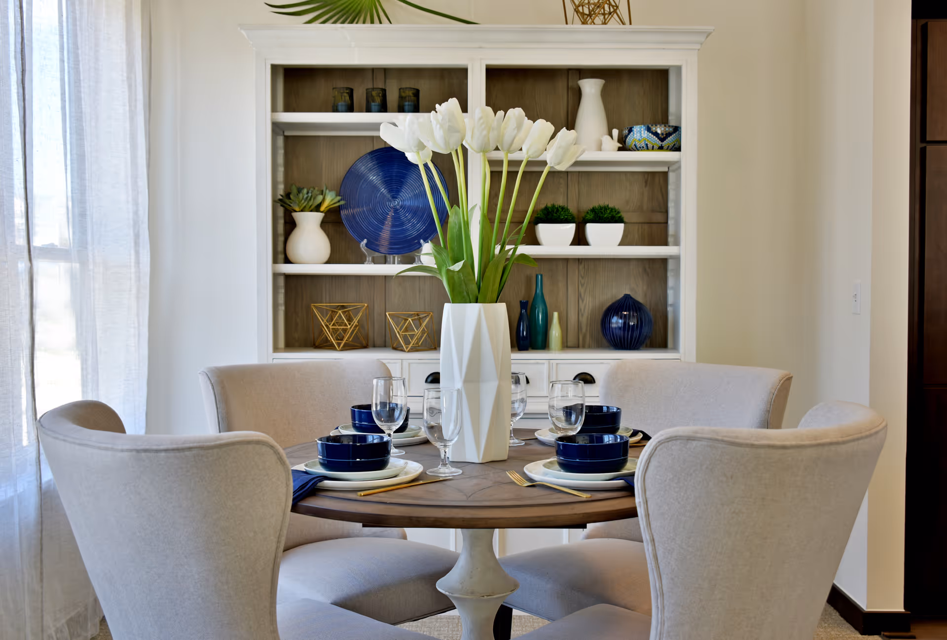 Round dining table set for four with a white geometric vase of tulips in front of a decorative hutch.