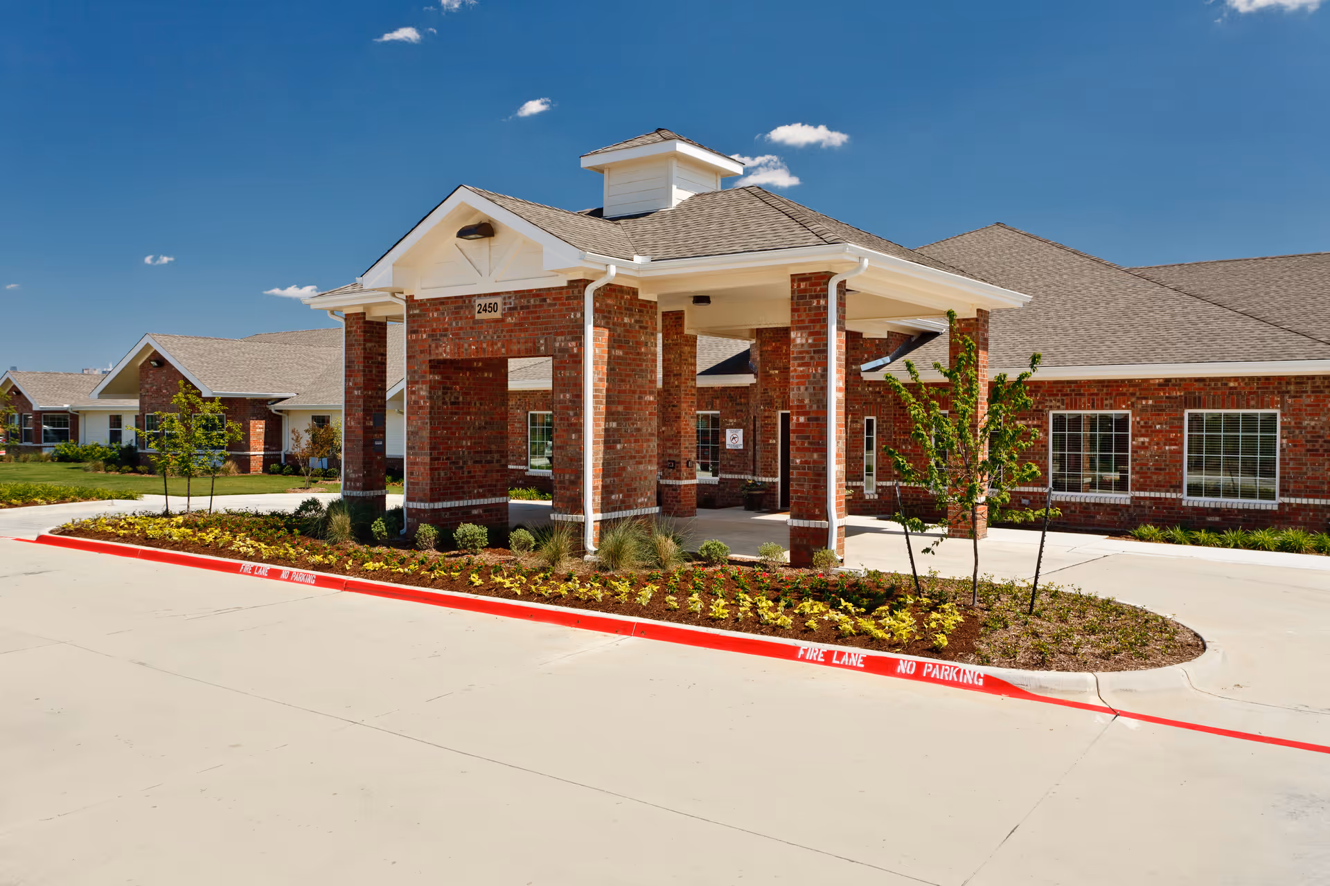 Front entrance of a red-brick senior care building with a covered porte-cochere, landscaped island, and clear blue sky.