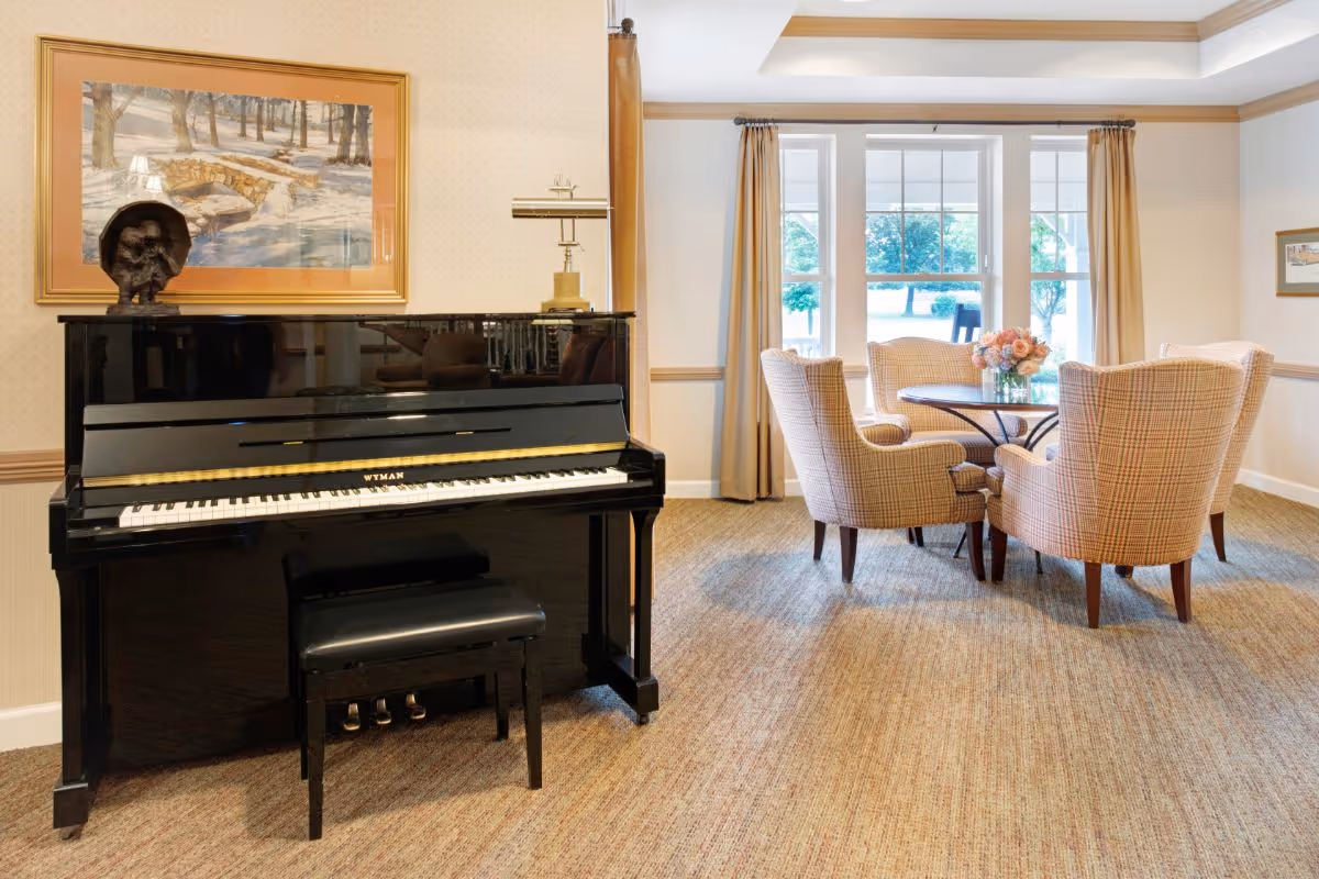 A bright common room with a black upright piano on the left and a round table surrounded by upholstered chairs by a large window.