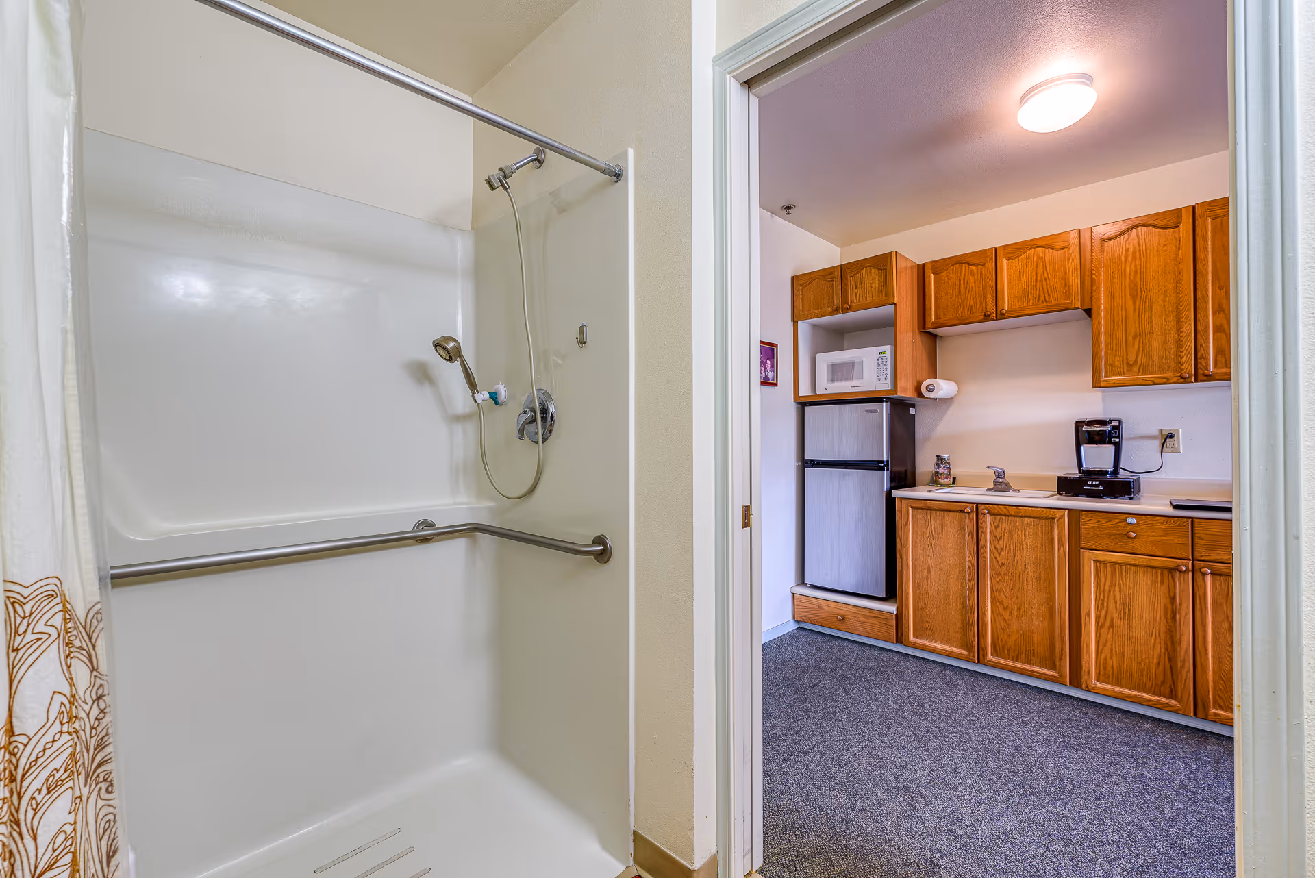 View of a bathroom shower area with a white shower curtain partially visible on the left, a handheld showerhead, and a safety grab bar. Through an open doorway, a small kitchen area is visible with wooden cabinets, a microwave, a mini refrigerator, a coffee maker, and a countertop with a paper towel holder.