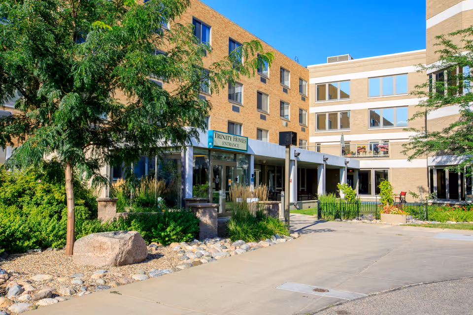 Front entrance of the Trinity Homes senior living building with brick facade, landscaped greenery, and a paved walkway.