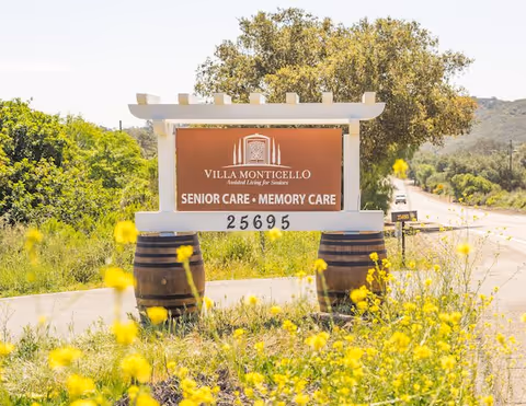 Outdoor view of a Villa Monticello sign for senior care and memory care, mounted on a white frame supported by two wooden barrels, surrounded by yellow wildflowers and greenery with a road and trees in the background.