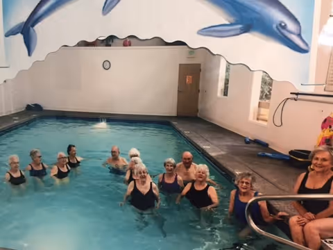 A group of elderly people participating in a water exercise class in an indoor swimming pool. The pool area has a mural of two dolphins on the wall above the pool. The participants are standing in the water, smiling and engaging in the activity.