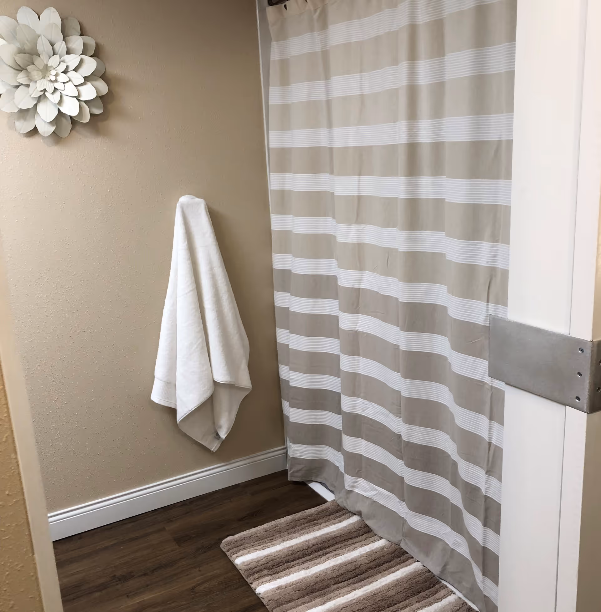 A bathroom corner featuring a beige and white striped shower curtain, a matching striped bath mat on a wooden floor, a white towel hanging on a beige wall, and a decorative white flower wall art.