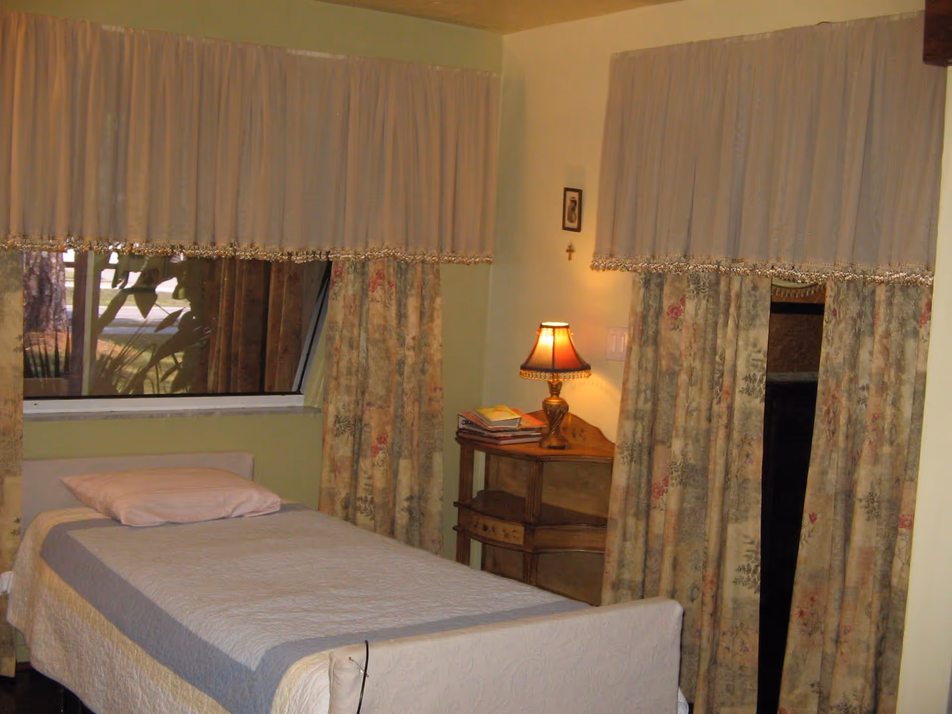 A small bedroom with a single bed covered with a white and gray quilt and a pink pillow. Next to the bed is a wooden nightstand with a decorative lamp and some books. The room has two windows with floral curtains and sheer white valances. The walls are painted light green.