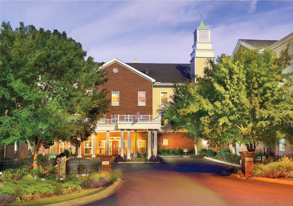Exterior view of Belmont Village Senior Living Green Hills building at dusk, featuring a brick and siding facade, a covered entrance with columns, an American flag, and surrounded by trees and landscaping.