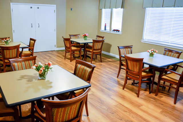 A bright dining room with several square tables, each surrounded by four wooden chairs with striped cushions. Small vases with flowers are placed on each table. The room has light-colored walls, large windows with blinds, and wood flooring.