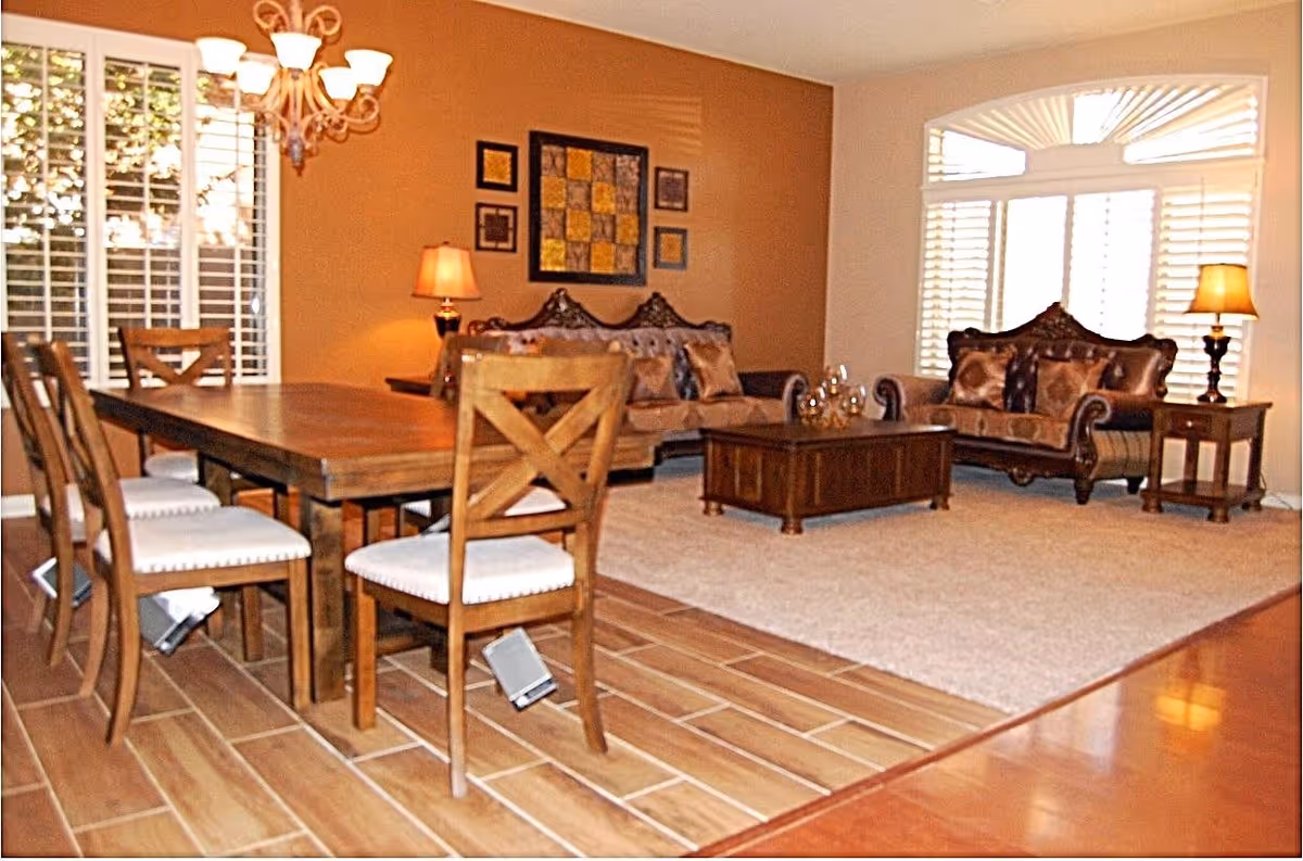A cozy living and dining area with wooden furniture. The dining area has a wooden table with six chairs featuring white cushions. The living area includes two ornate sofas with patterned cushions, a wooden coffee table, and two side tables with lamps. The room has large windows with white shutters, a beige carpet, and warm lighting from a chandelier and lamps.