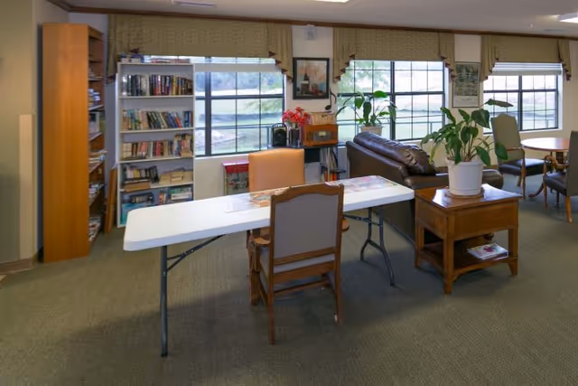 A cozy common area with large windows letting in natural light. The room features a white folding table with chairs, a wooden bookshelf filled with books, a brown leather couch, a wooden side table with a potted plant, and additional seating around a round table near the windows.