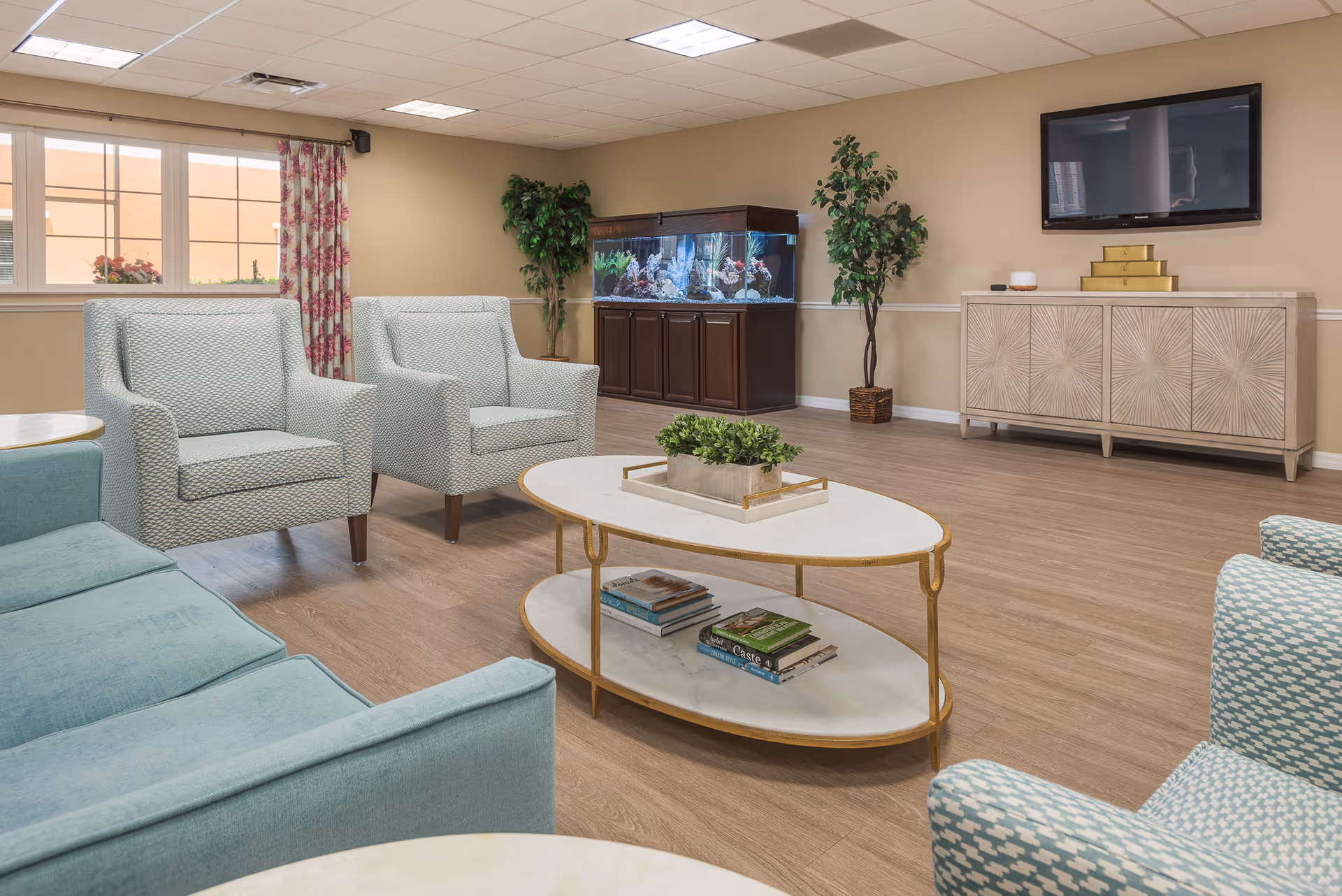 A bright and spacious living room area with light blue upholstered chairs and a sofa arranged around an oval coffee table with books and a small plant on it. There is a large fish tank on a wooden cabinet against the beige wall, flanked by two potted plants. A flat-screen TV is mounted on the wall above a decorative sideboard. Large windows with floral curtains allow natural light into the room.