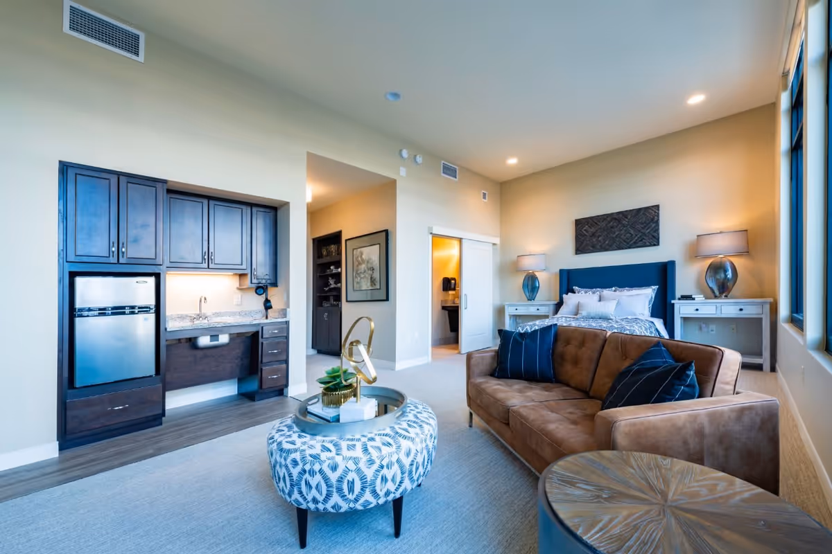 A spacious and well-lit studio apartment featuring a brown sofa with dark blue pillows, a patterned round ottoman with a decorative tray, a bed with white and blue bedding, two bedside tables with lamps, a kitchenette with dark wood cabinets, a small refrigerator, and a sink. The room has large windows allowing natural light to fill the space, beige walls, and light carpet flooring.