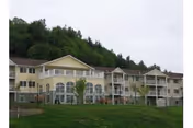 Exterior view of a multi-story senior living facility building with balconies and large windows, surrounded by green grass and trees in the background under a cloudy sky.