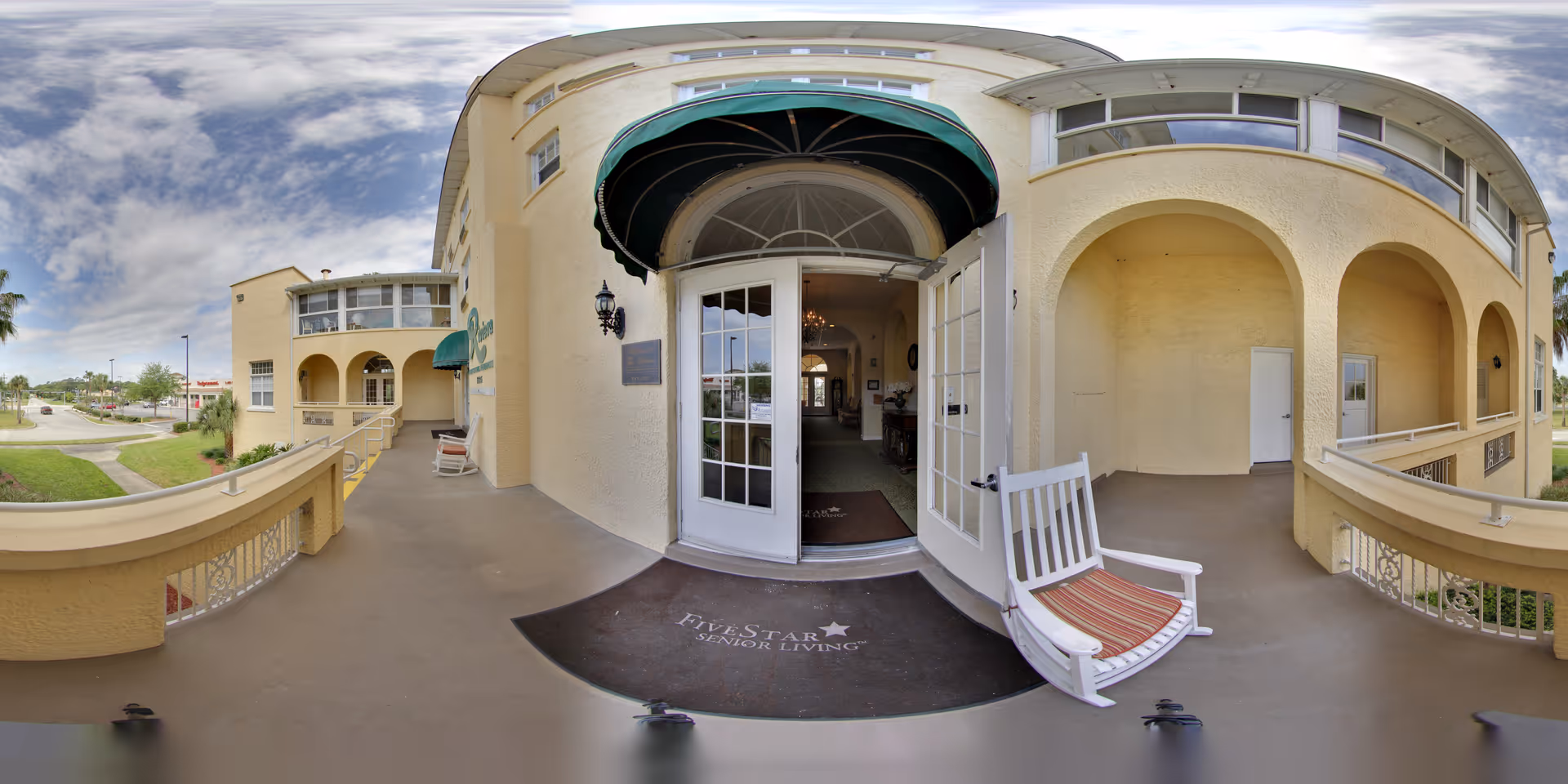 Wide-angle view of the entrance to a senior living facility with beige walls and arched architectural features. The entrance has double glass doors, one of which is open, leading inside. A green awning is above the door, and a white rocking chair with a striped cushion sits on the right side of the entrance. A dark mat with the text 'Five Star Senior Living' is placed in front of the door. The exterior area includes a balcony with railings and a view of a street and greenery in the background under a partly cloudy sky.