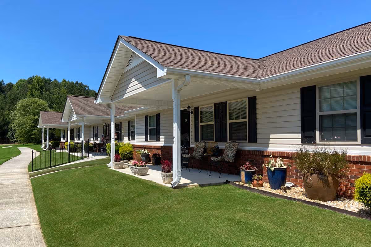 Single-story senior living building front with covered porches, potted plants, and a sidewalk beside a green lawn under a clear blue sky.