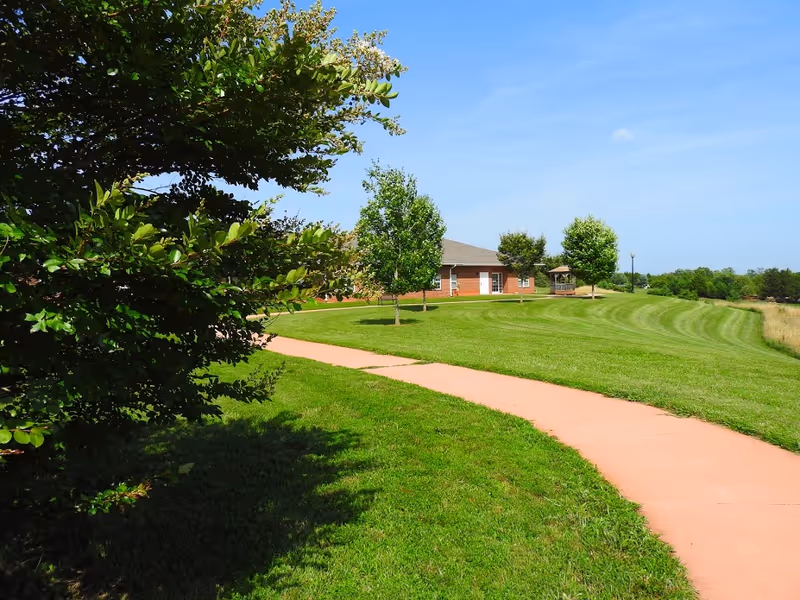 A paved walkway curves through a well-maintained grassy area with several small trees and bushes. In the background, there is a single-story brick building with a gazebo nearby under a clear blue sky.