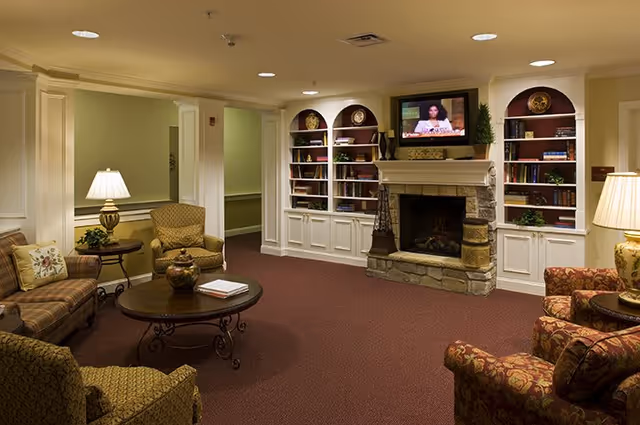 A cozy living room area in a senior living facility featuring a stone fireplace with a TV mounted above it, built-in white bookshelves on either side filled with books and decorative items, multiple upholstered armchairs and sofas arranged around a round coffee table, and warm lighting from table lamps.