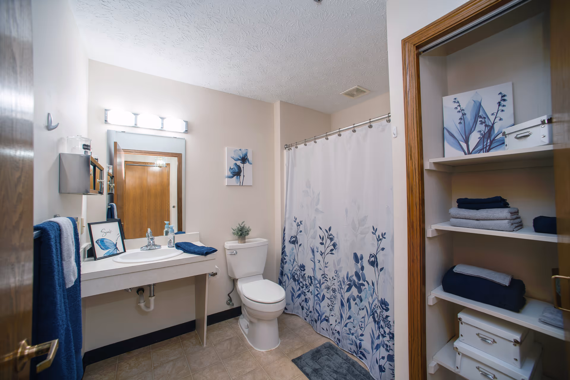 Clean, well-lit bathroom with a sink and mirror on the left, a toilet and floral shower curtain in the center, and open shelving with towels on the right.