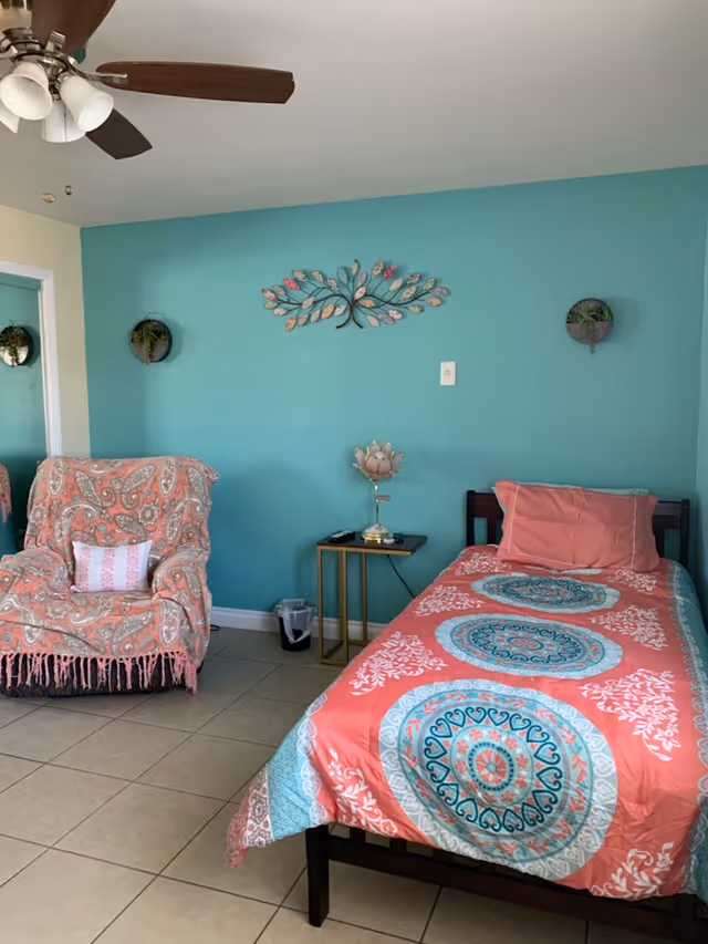Twin bed with coral and teal patterned bedding next to an armchair and side table against a turquoise accent wall decorated with metal leaf art.